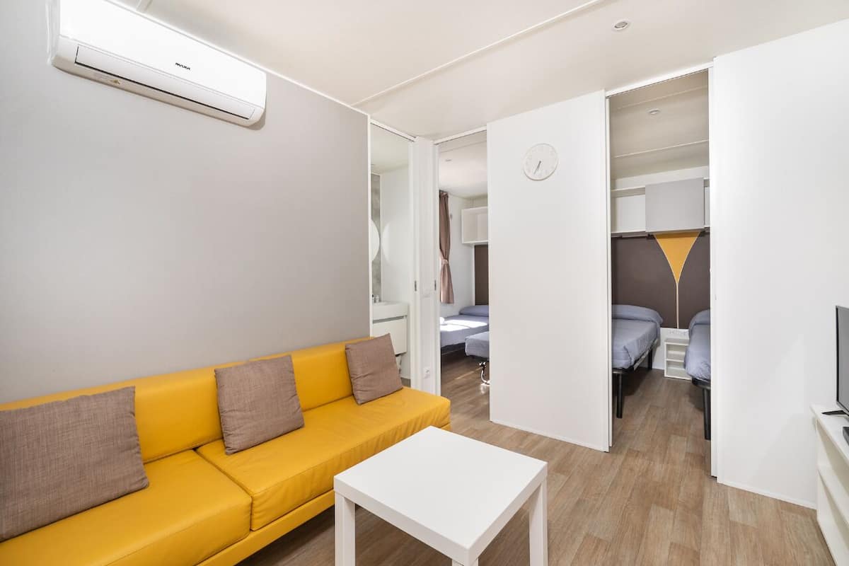 Ein modernes, helles Zimmer mit einem gelben Sofa, einem weißen Tisch und einem Bett im Hintergrund.