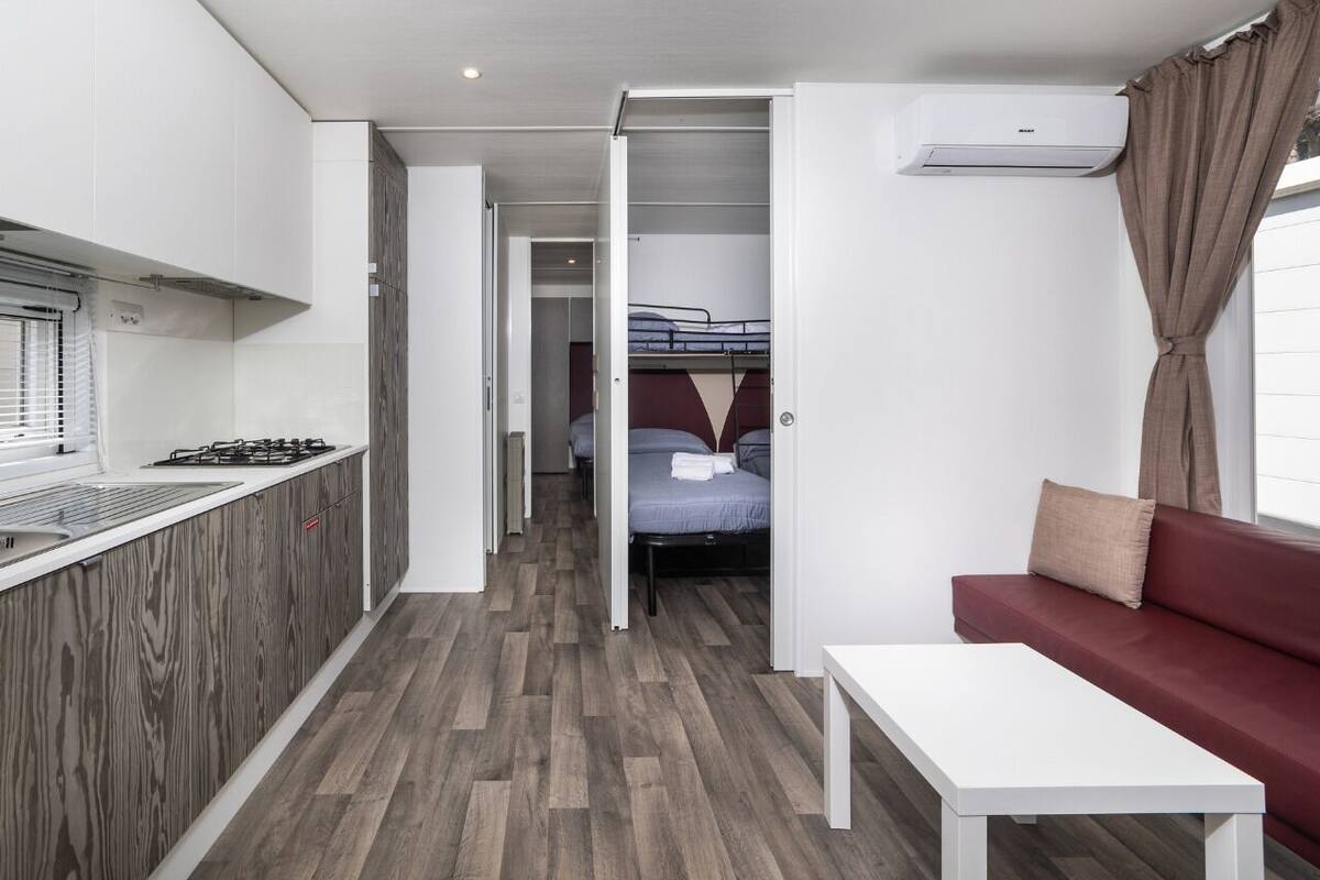 Ein kleines Hotelzimmer mit einer Küchenzeile, einem Bett mit Kopfteil, einem roten Sofa und einem Fenster mit Vorhängen.