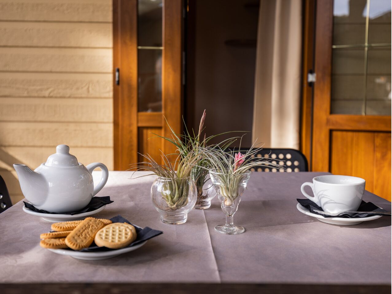 A table set with a white teapot, two glass vases with plants, a plate of cookies, and a white cup on a table with a view of a wooden door and window.