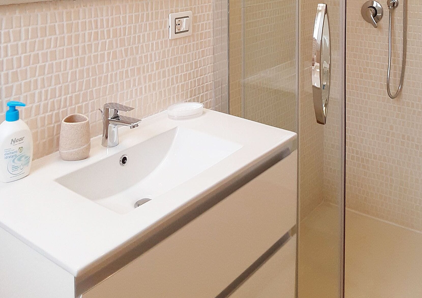 A bathroom with a white sink, a mirror, a glass shower enclosure, and a beige tiled wall.