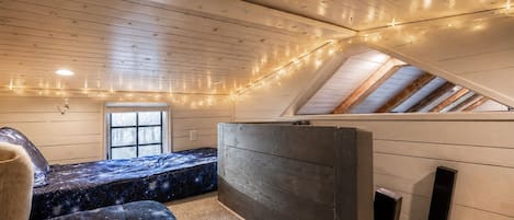 Un dormitorio acogedor en el ático con techo inclinado, una cama con manta de diseño galáctico, un ventanal pequeño y luces colgantes.