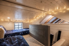 Une chambre mansardée chaleureuse, avec un plafond en pente, un lit recouvert d’une couette sur le thème de la galaxie, une petite fenêtre et des guirlandes lumineuses.