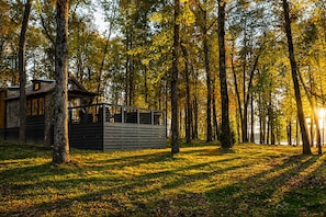 Une cabane dotée d’un toit en pente, entourée d’une forêt où s’élèvent de grands arbres, et bordée d’une zone herbeuse.