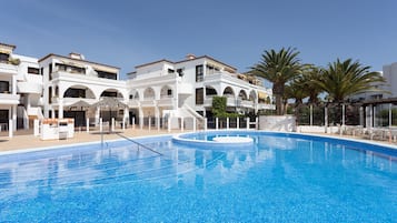 Una piscina con superficie de agua azul cristalina, rodeada de edificios blancos con balcones y una palmera.