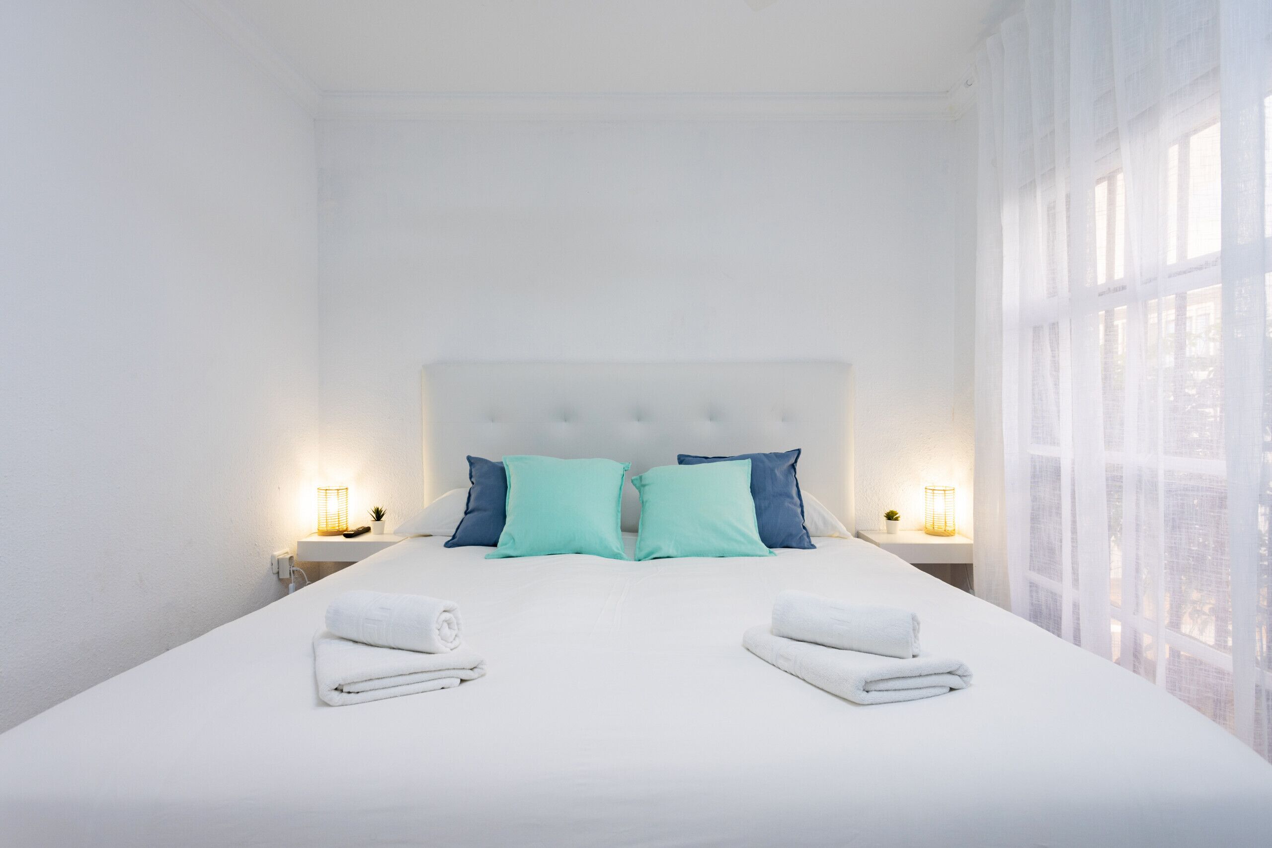 Habitación de hotel con una cama grande, dos almohadas, dos mesitas de noche con lámparas y una ventana con cortinas translúcidas.
