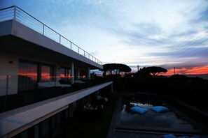Un edificio moderno con balcón que da a una piscina reflectante y vistas al atardecer.