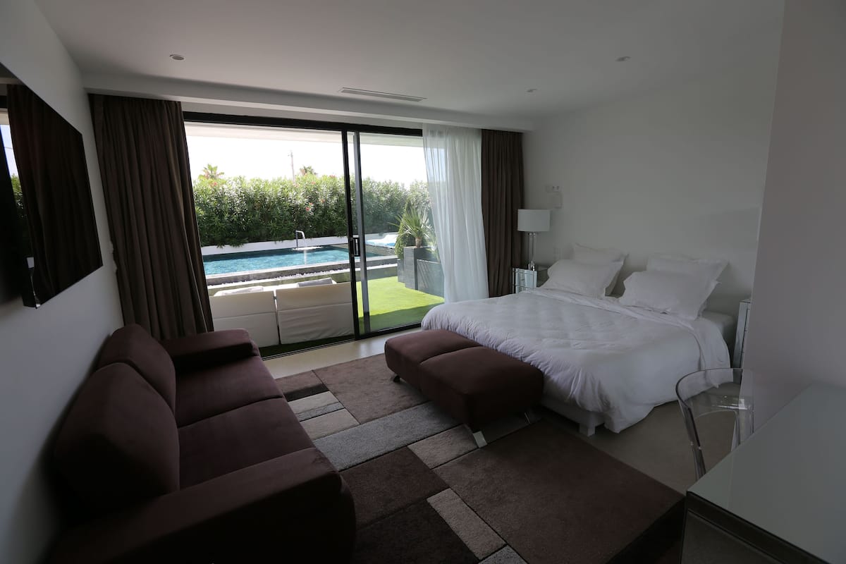 A modern hotel room with a large bed, a sofa, a glass door leading to a pool, and a view of greenery.