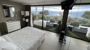 A modern bedroom with a large bed, a bathroom with a vanity, and a balcony with a view of the sea. - Villa Miami (Roquebrune-sur-Argens)