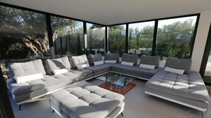 A modern living room with a sectional sofa, a glass coffee table, and large windows offering a view of greenery. - Villa Miami (Roquebrune-sur-Argens)