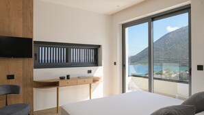 A modern bedroom with a large bed, a desk with a TV, and a balcony with a mountain view. - Nandana Luxury Villa, an infinite blue experience (Rethymno)