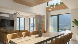 A modern living room with a large wooden dining table, comfortable seating, a flat-screen TV, and a view of the sea through large windows. - Nandana Luxury Villa, an infinite blue experience (Rethymno)