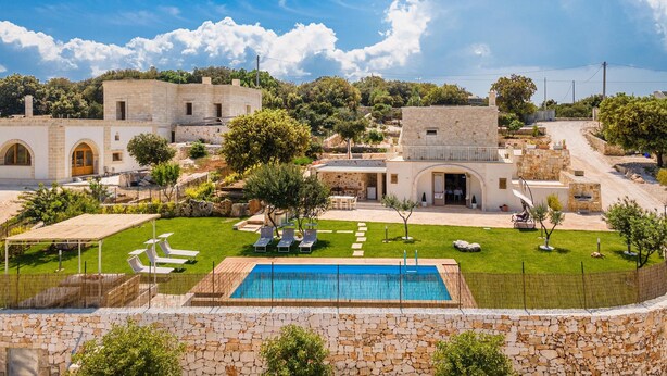 A pool area with a stone wall, a building with a stone facade, and a clear blue sky. - 2943 Casa Marangi by Barbarhouse (Ostuni)
