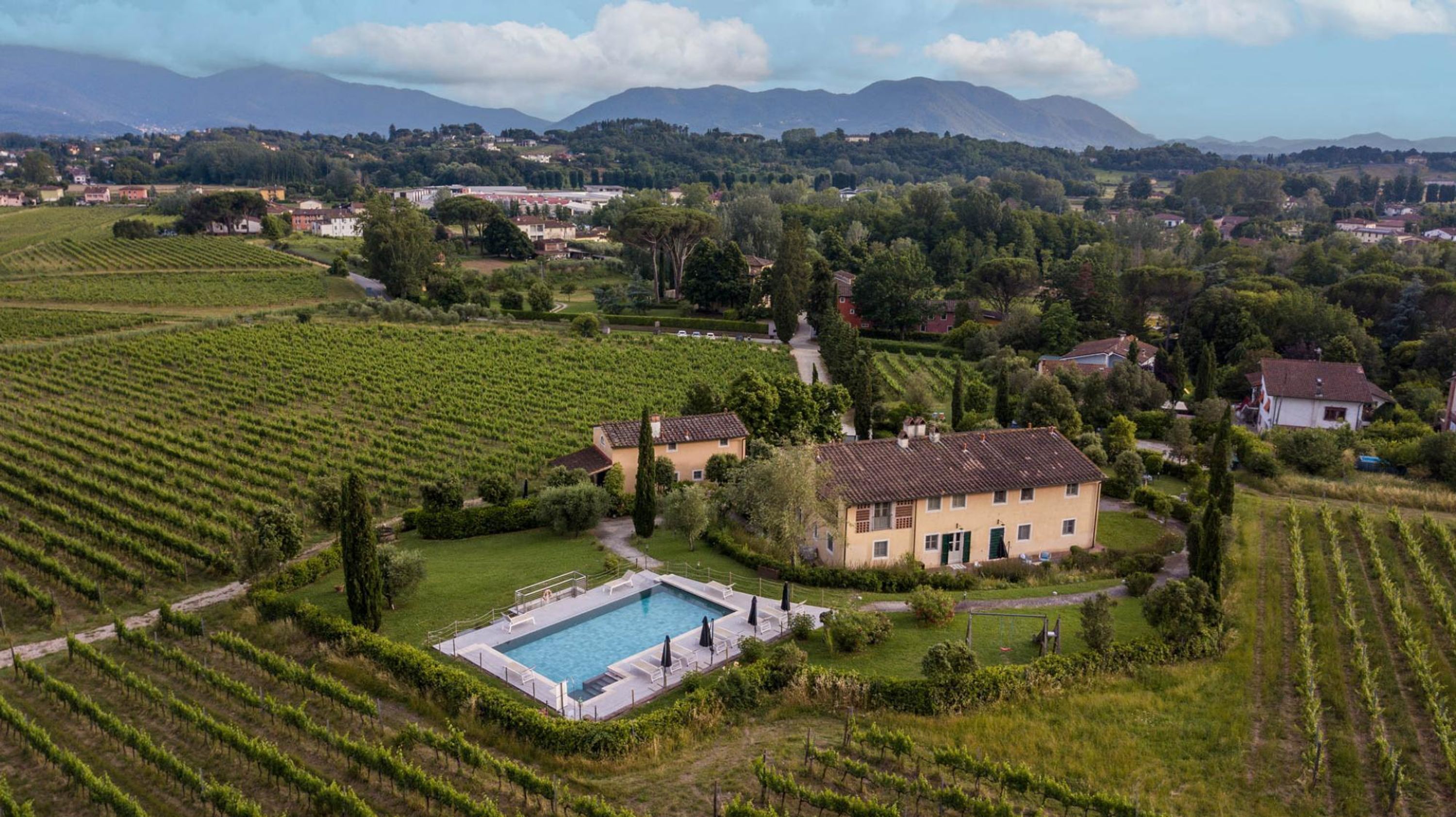 Uma villa com piscina, rodeada por fileiras de vinhas, inserida numa paisagem montanhosa com montanhas ao longe.