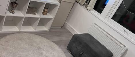 Una sala de estar con un sofá gris, un mueble blanco para el equipo de entretenimiento con un televisor, un radiador blanco y una ventana con cortinas.