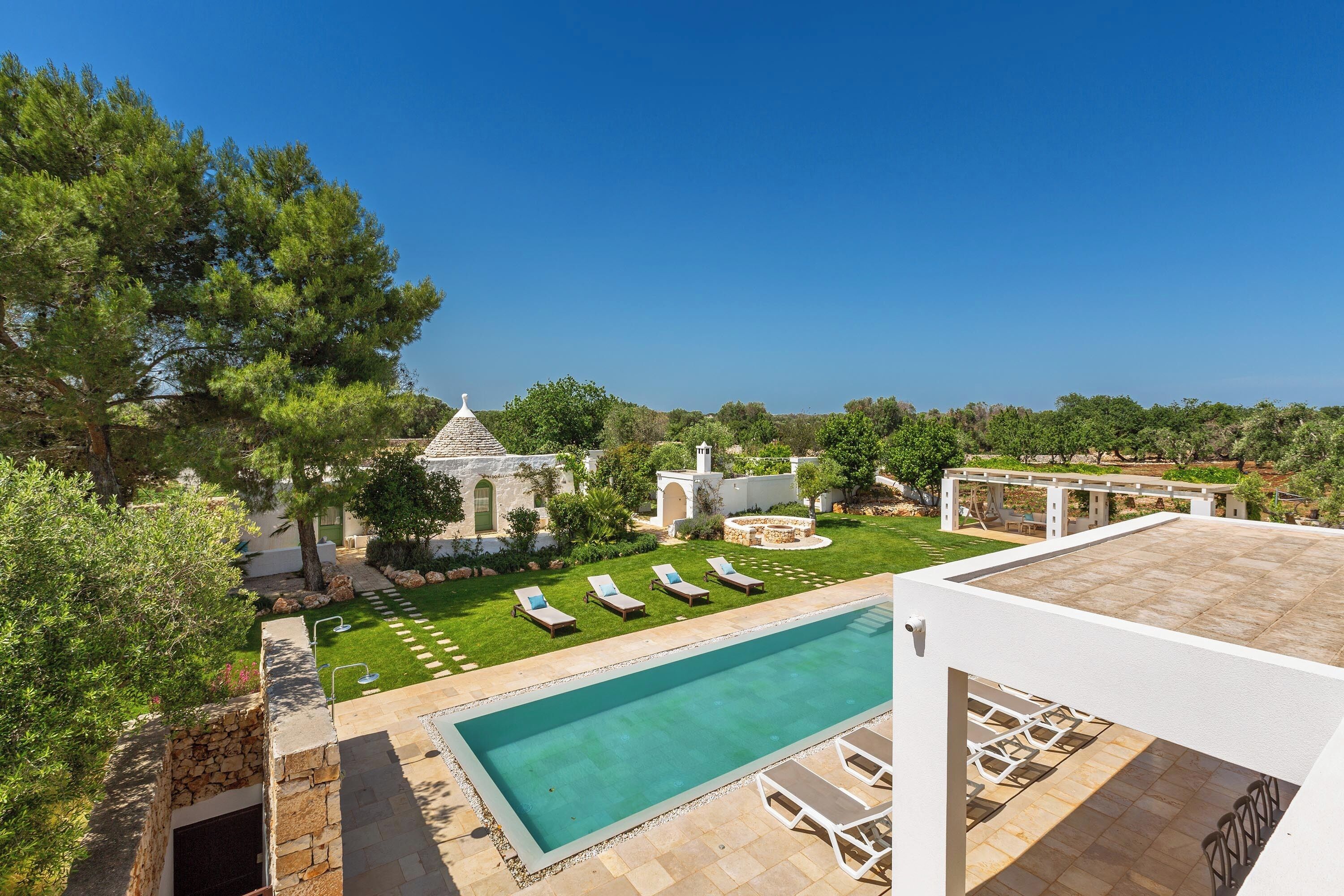 A swimming pool with lounge chairs, a stone wall, and a view of a garden and buildings.