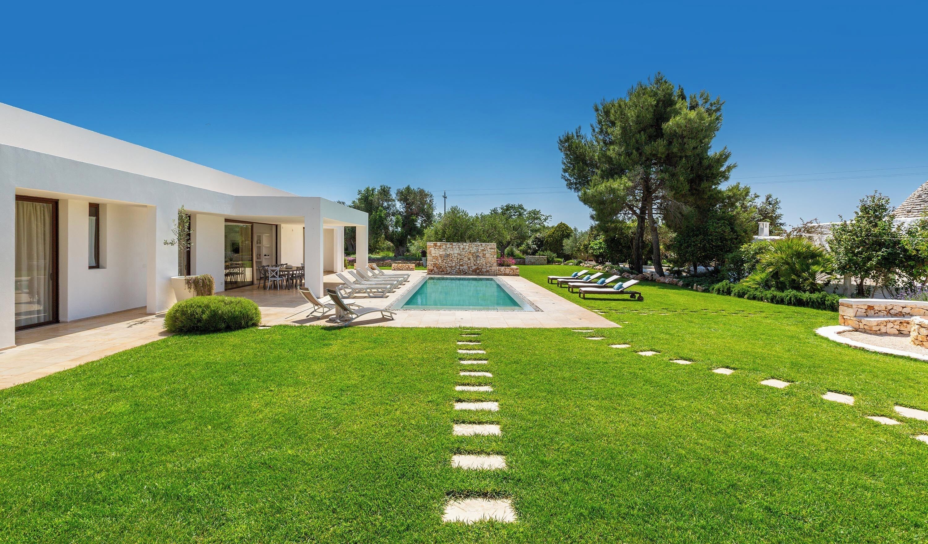 Una casa moderna con piscina, un patio con sillones de descanso y un césped cuidado.