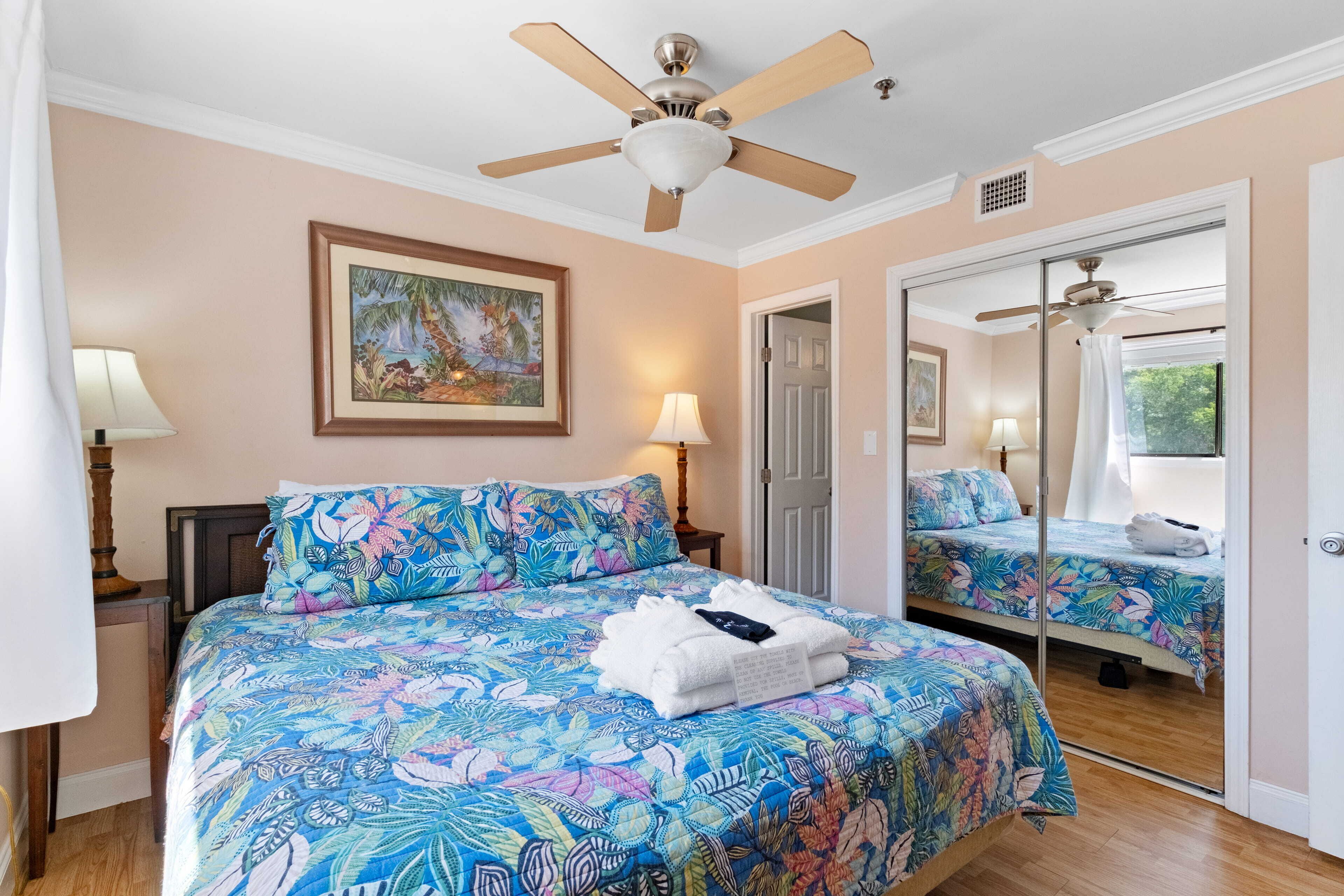 A bedroom with a large bed, a ceiling fan, a mirror, and a door leading to another room.