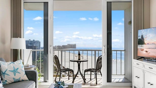 En balkon med et bord og stole, en sofa, et fjernsyn og udsigt til både strand og bybillede.