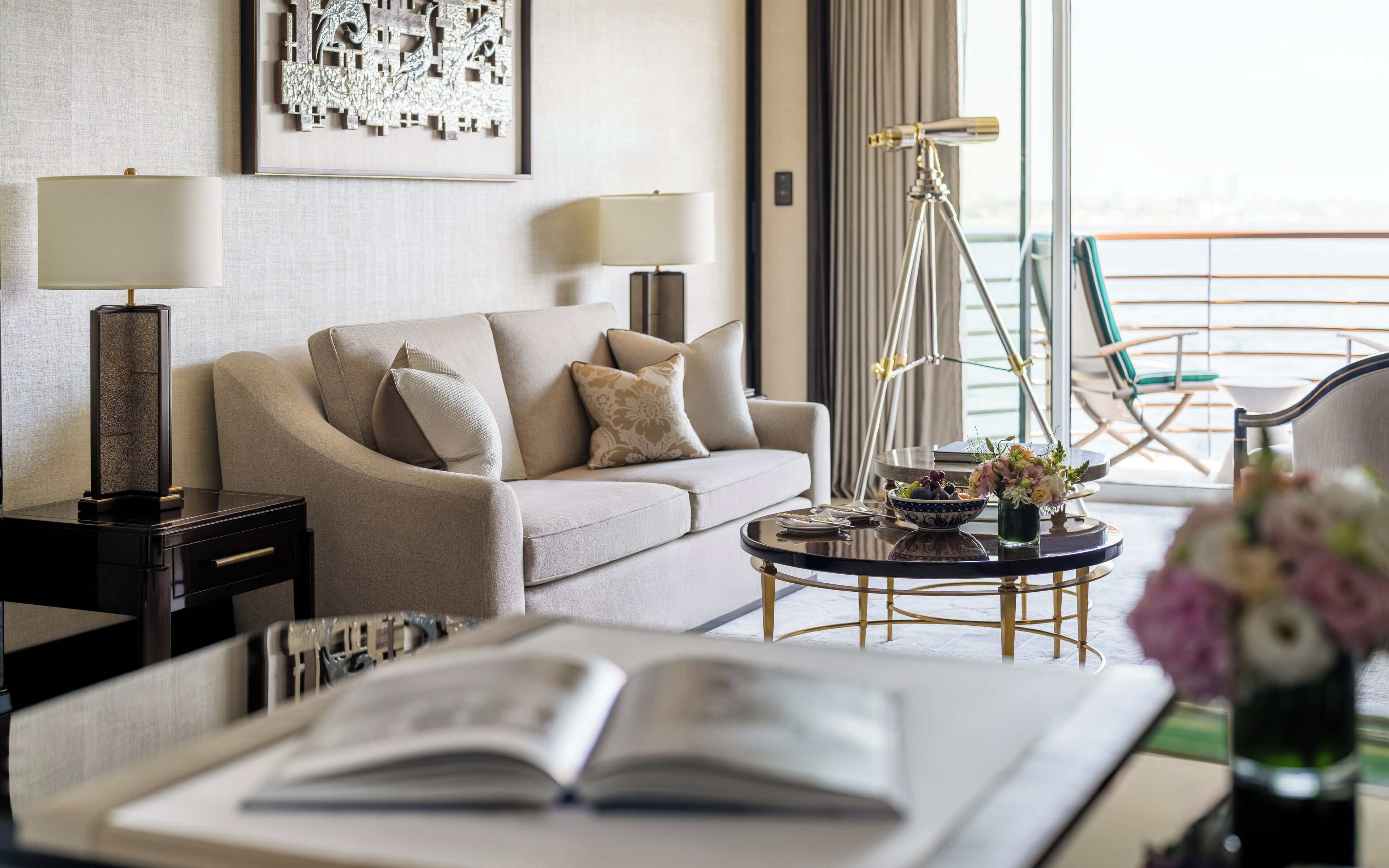 Una habitación de hotel moderna con sofá, dos lámparas, una mesita auxiliar, una mesa de centro con un cuenco y flores, y un balcón con sillas.