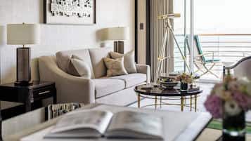 Una habitación de hotel moderna con sofá, dos lámparas, una mesita auxiliar, una mesa de centro con un cuenco y flores, y un balcón con sillas.