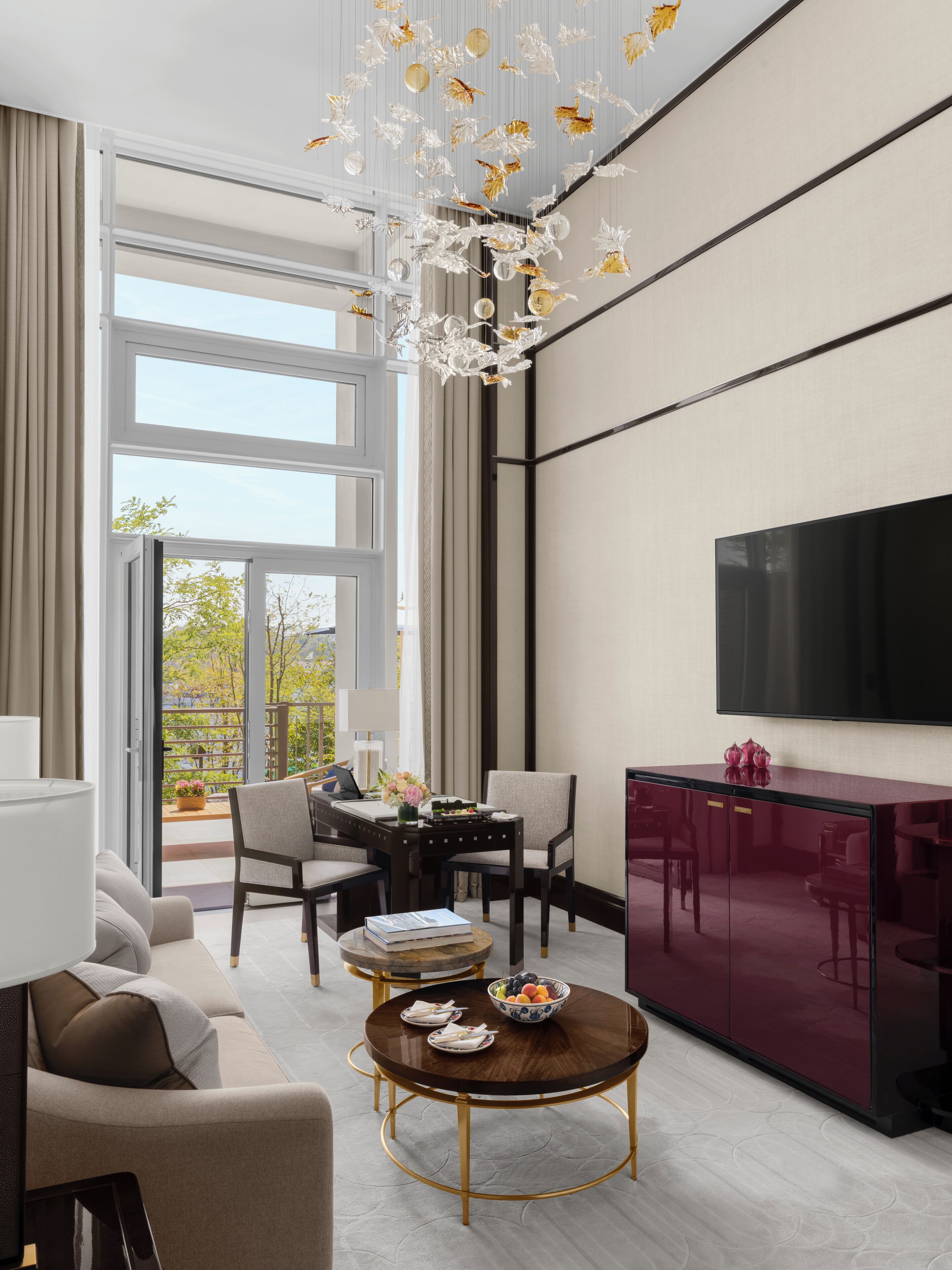 A modern living room with a sofa, coffee table, dining table, and chairs. There is a TV mounted on the wall and a balcony with a view of trees.