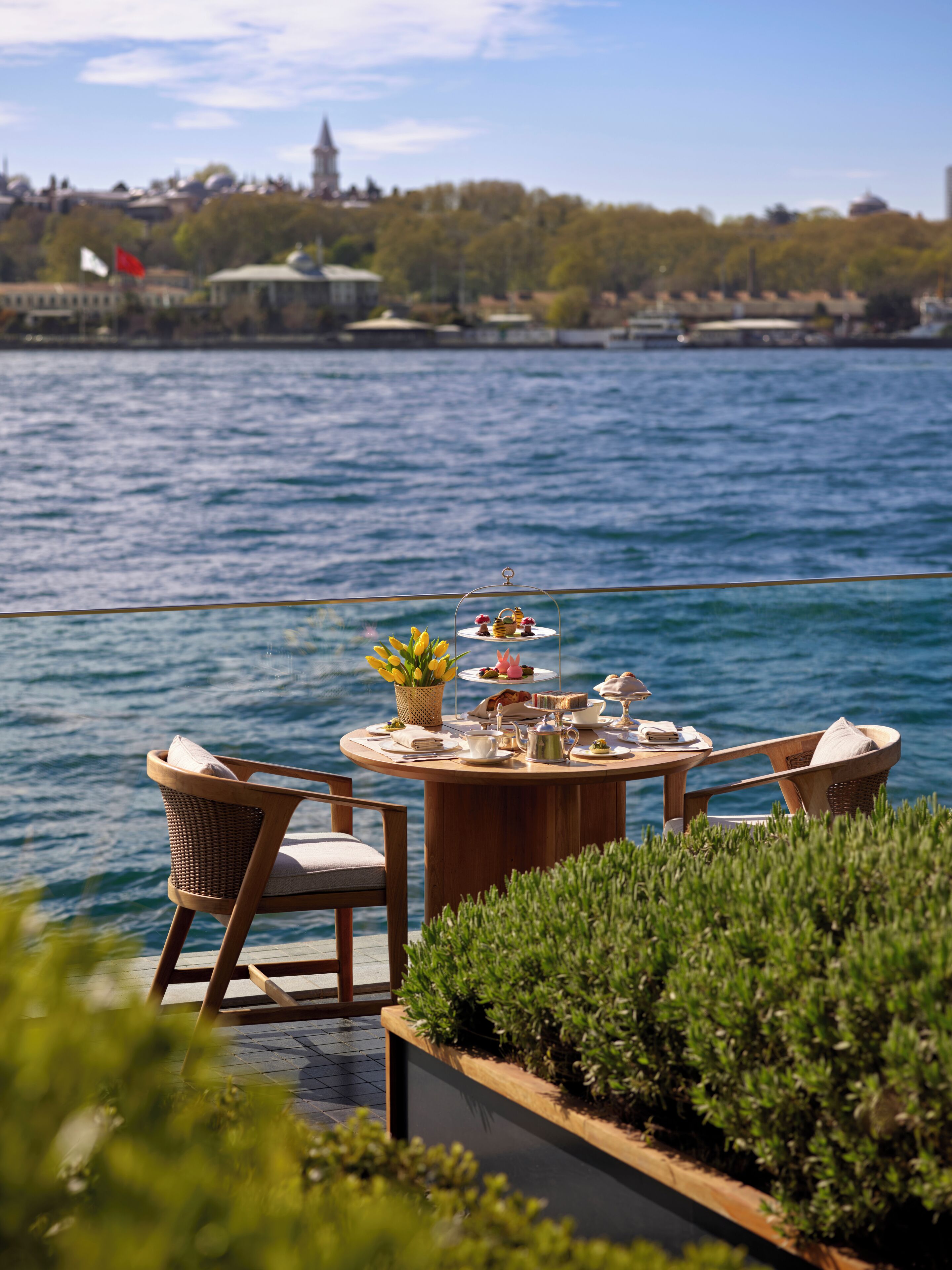 A round wooden table with chairs, a small garden, and a view of a body of water with buildings in the background.