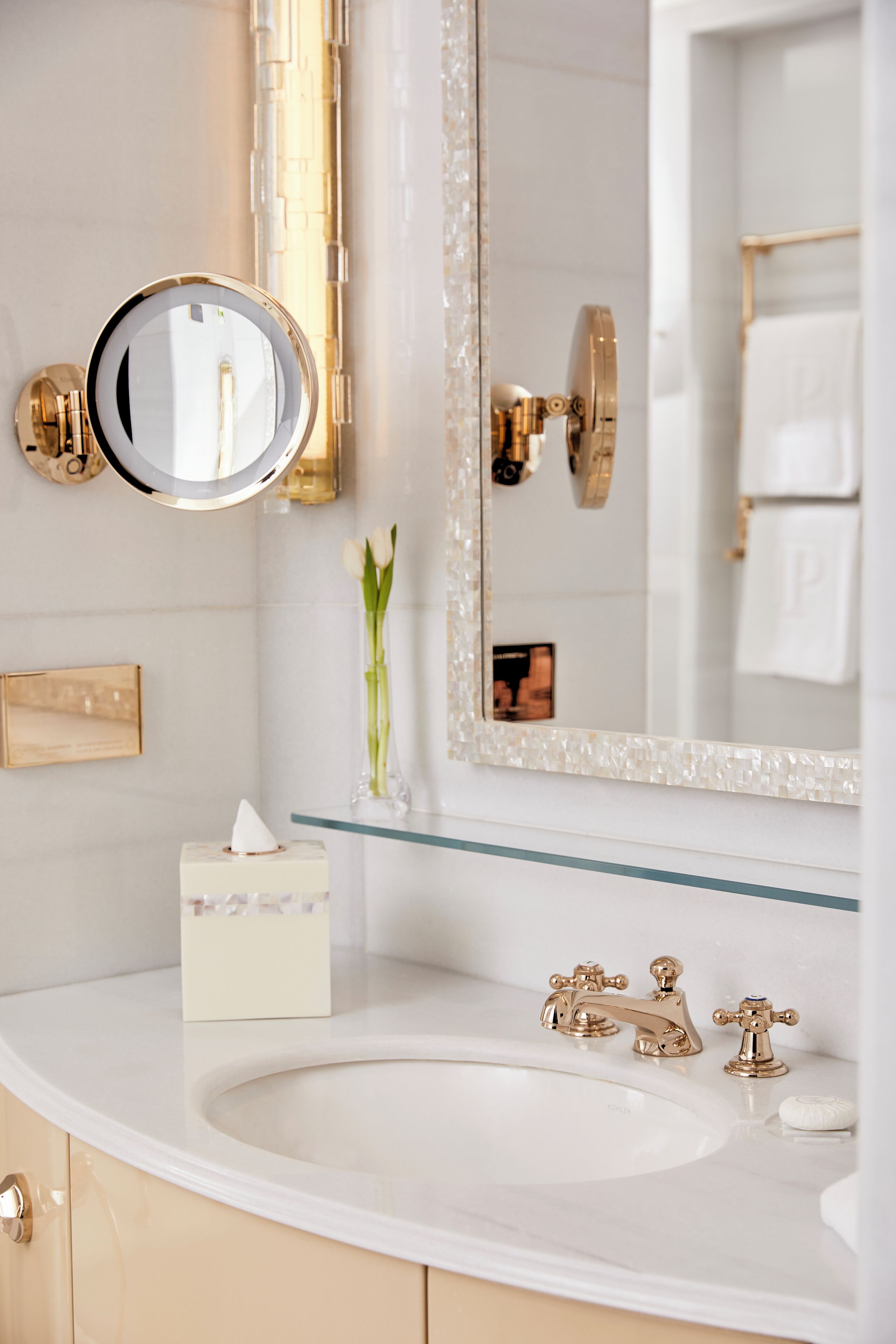 A bathroom with a white sink, gold faucets, a round mirror with a gold frame, and a vase with white flowers on the counter.