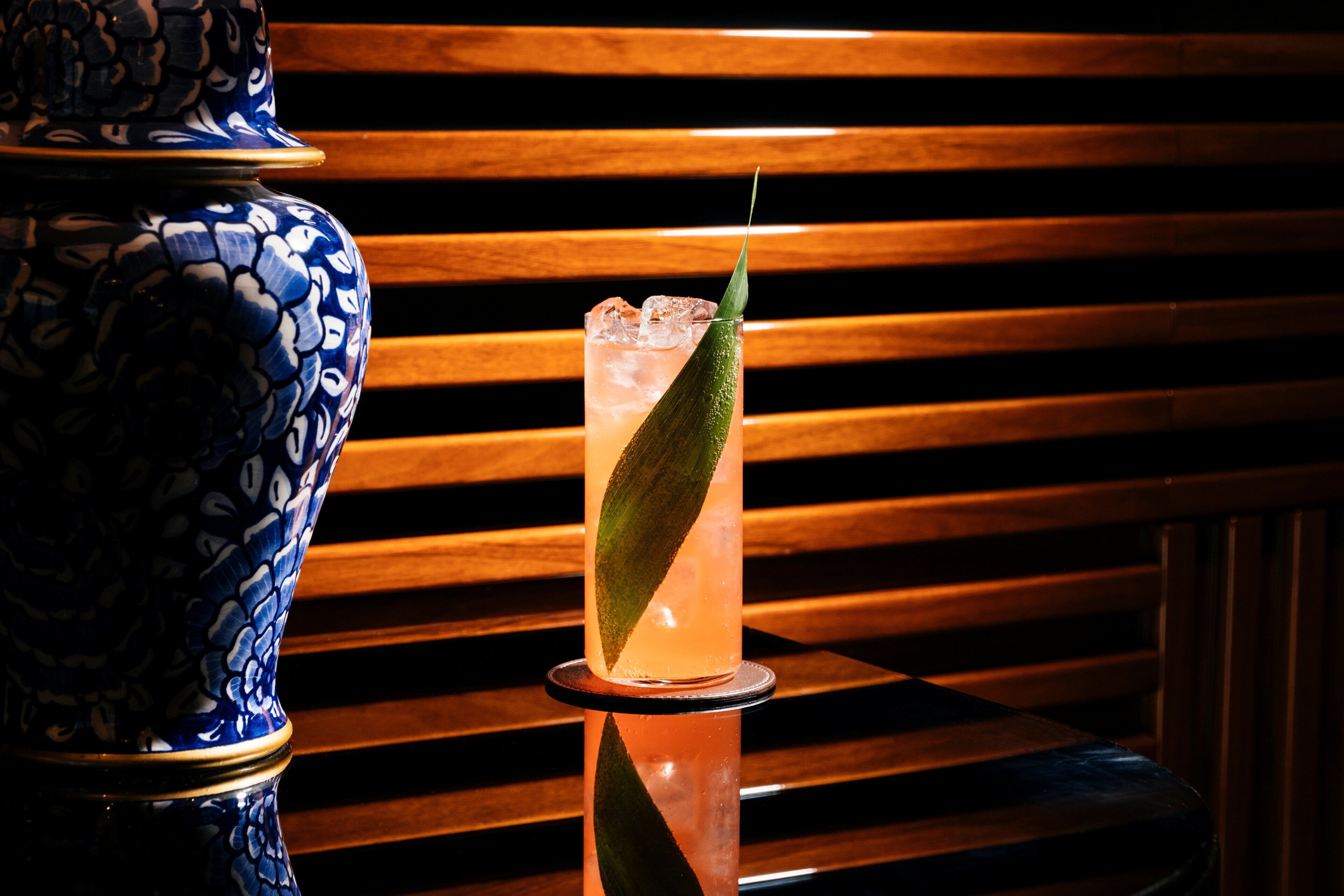 A tall glass with a green leaf garnish, ice cubes, and a reflective base, placed on a reflective surface with a blue and white patterned vase in the background.