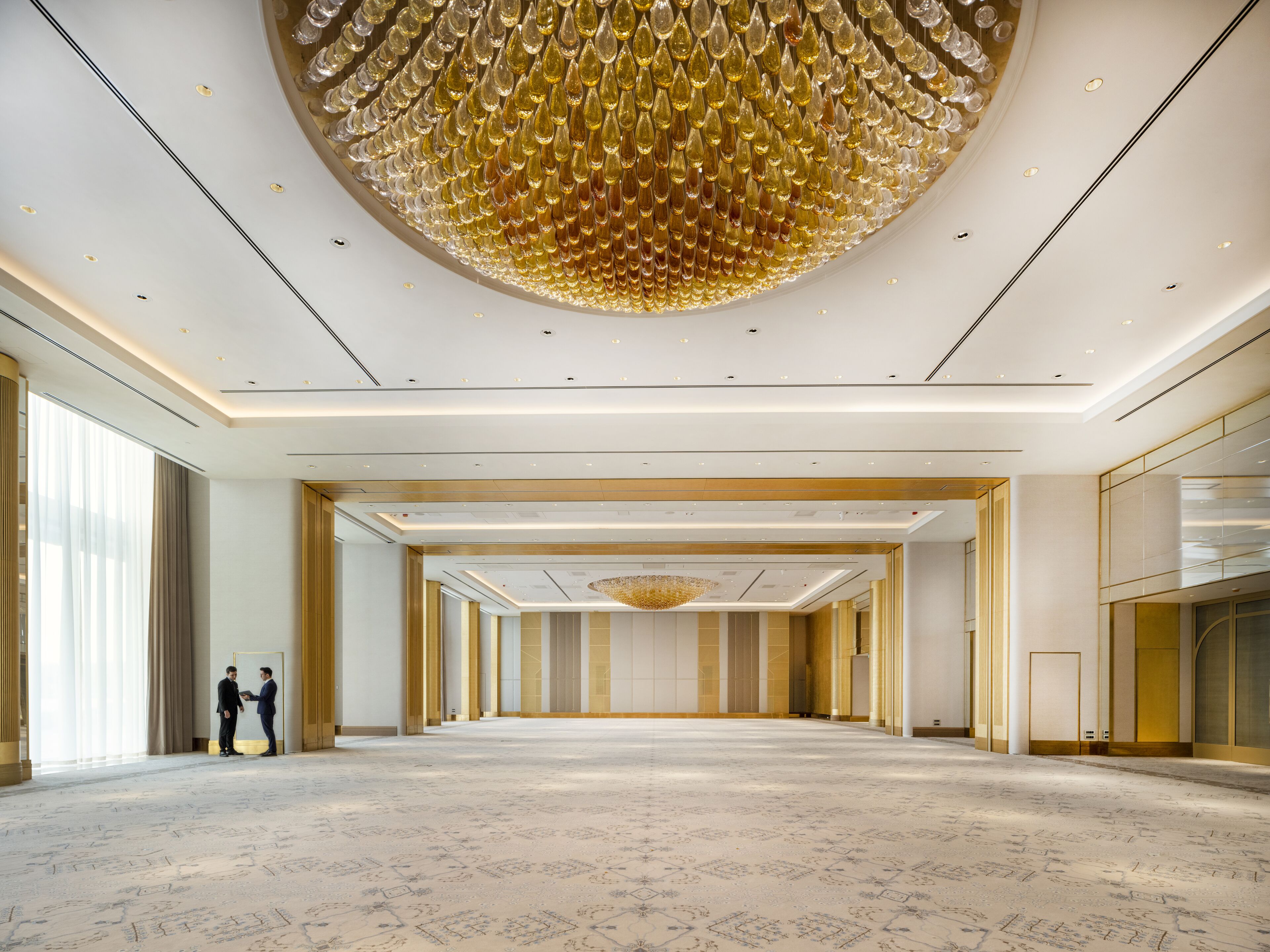 A spacious, well-lit hotel lobby with a large chandelier, high ceilings, and a patterned carpet.