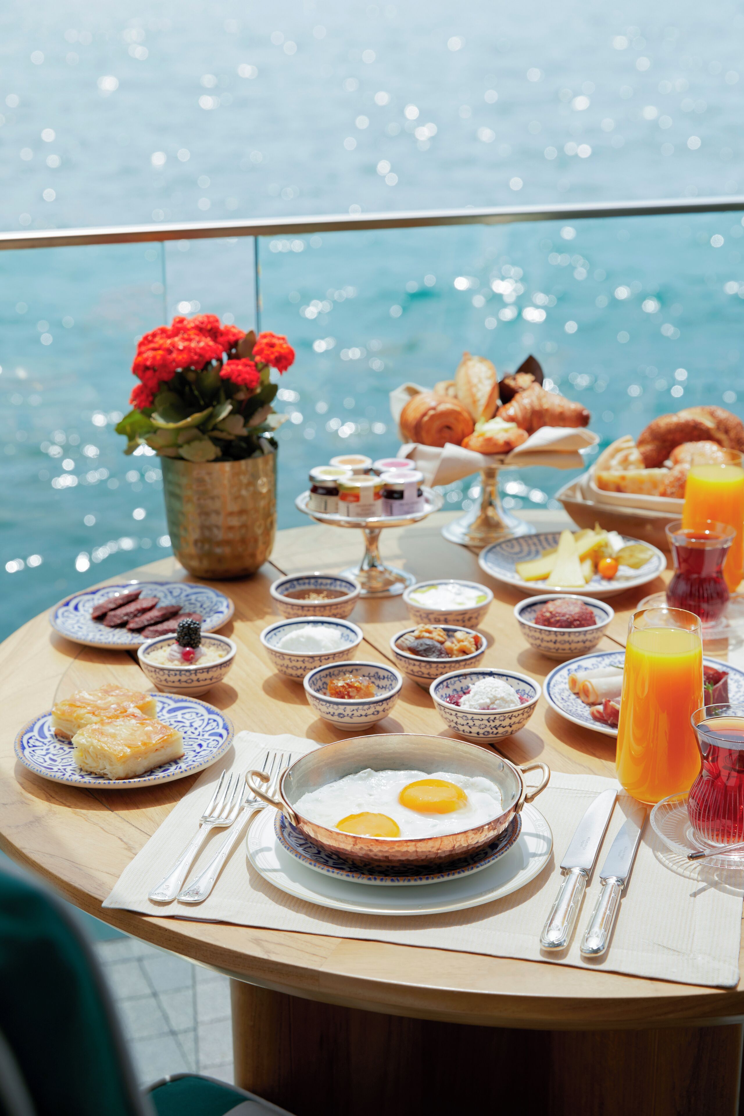 A breakfast table with various dishes, a vase of flowers, and a view of the sea.
