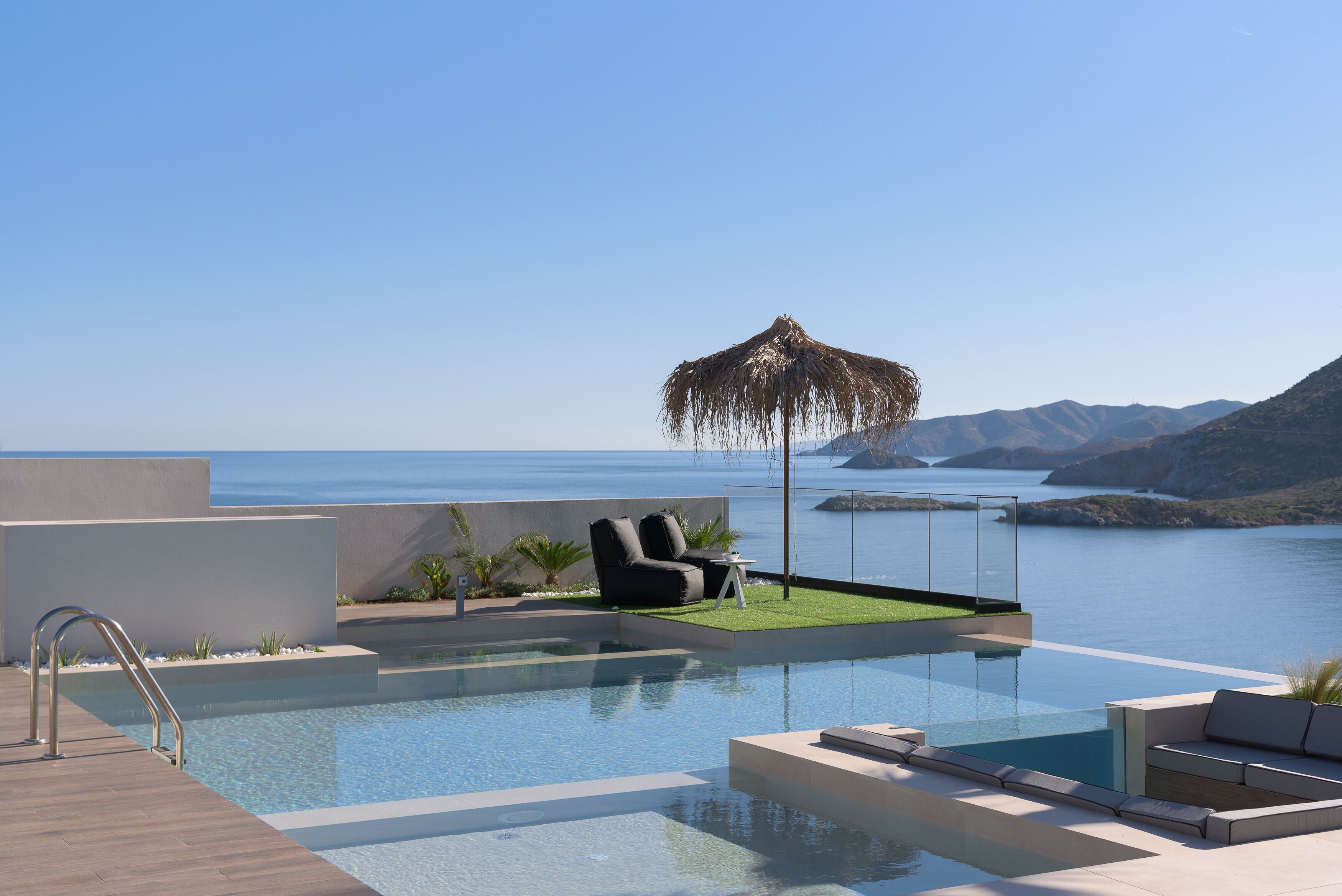 A modern infinity pool with a clear view of the sea and mountains, a thatched umbrella, and contemporary outdoor seating.