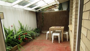 Een klein, afgesloten terras met een houten tafel en witte plastic stoelen, omringd door potplanten en een tegelvloer.