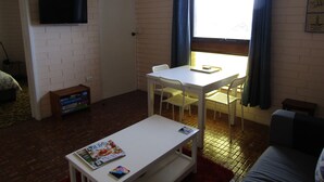 Een kleine, goed verlichte kamer met een witte tafel, twee stoelen, een aan de muur bevestigde televisie en een raam met gordijnen.