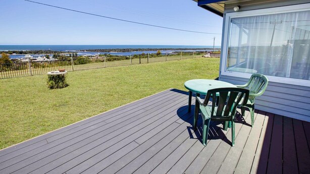 A deck with a table and chairs, overlooking a grassy area with a view of the sea and a fenced property. - Kalimna Holiday House: Relax and enjoy the magnificent 180-degree water views (Kalimna)