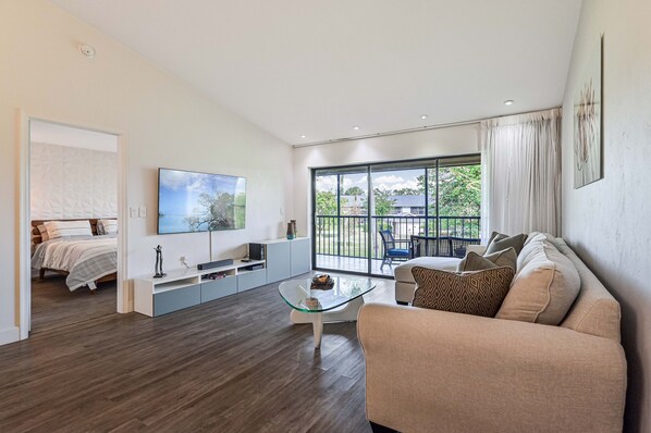A modern living room with a glass coffee table, a sofa, a TV, and a balcony with a view of greenery.