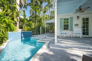 Une piscine dont le bord est revêtu de carrelage, entourée de palmiers et dotée d’un patio couvert avec des meubles blancs.
