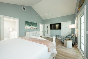 A bedroom with a large bed, a ceiling fan, a painting, a TV, and a chair.