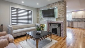 Una sala de estar con sofá, mesa de centro, chimenea y una zona de cocina con armarios y televisor montado en una pared de piedra.