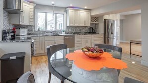 Una cocina moderna con gabinetes blancos, un refrigerador de acero inoxidable y una zona de comedor con una mesa de vidrio y un mantel individual naranja.