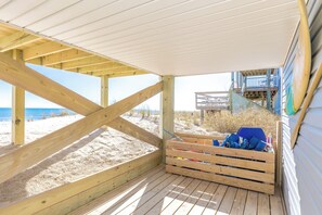 Una terraza de madera con vistas a la playa y al océano, un área de descanso cubierta con cojines azules, y una estructura de madera con barandilla azul.