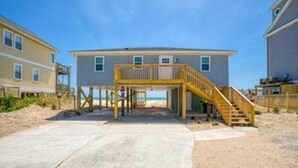 Une maison de plage à deux étages, dotée d’une terrasse en bois et d’un escalier menant à un niveau supérieur.