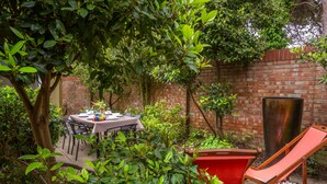 Un patio con mesa y sillas, rodeado de follaje y un muro de ladrillo.