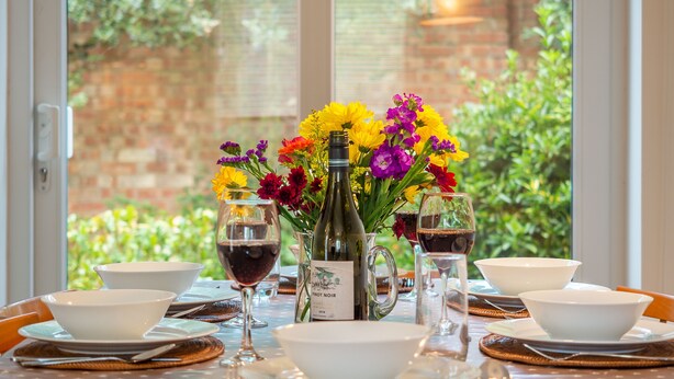 Mesa lista para una comida con cuencos blancos, copas de vino y una botella de vino, con vista a la vegetación por la ventana.