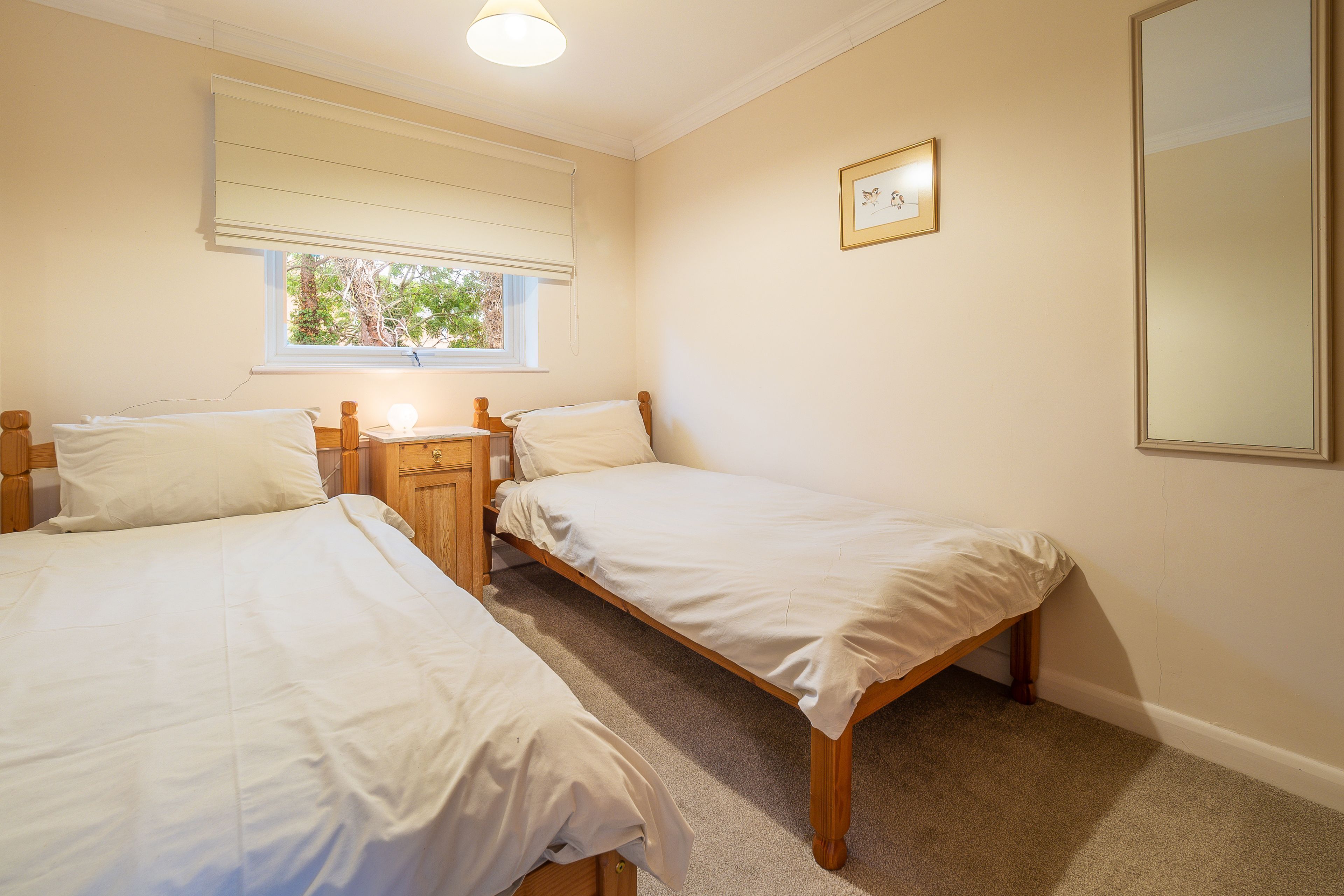 A bedroom with two single beds, a wooden nightstand, a bedside table, a window with blinds, and a framed picture on the wall.