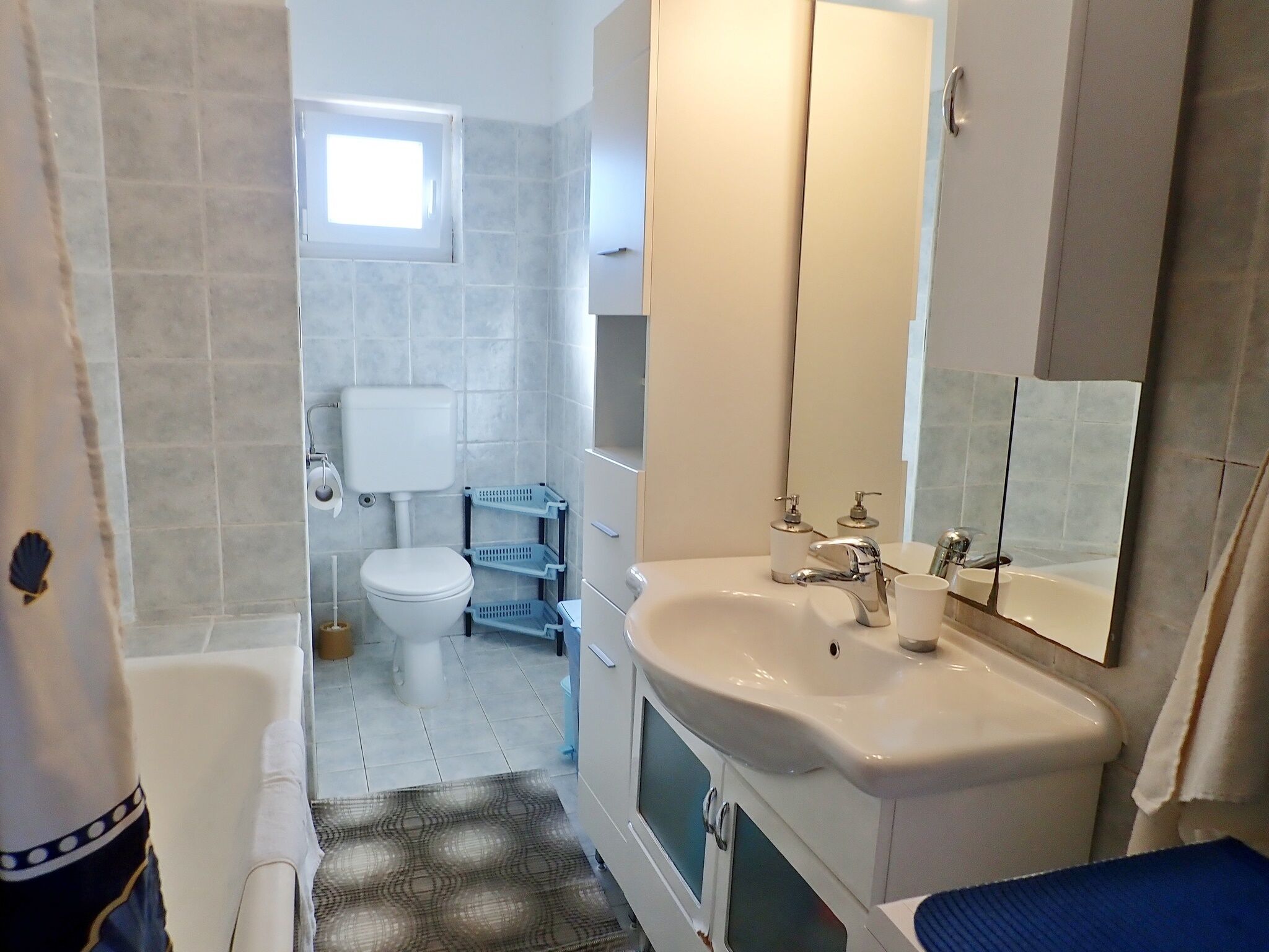A bathroom with a toilet, sink, and mirror. There is a window above the toilet and a shelf with toiletries.