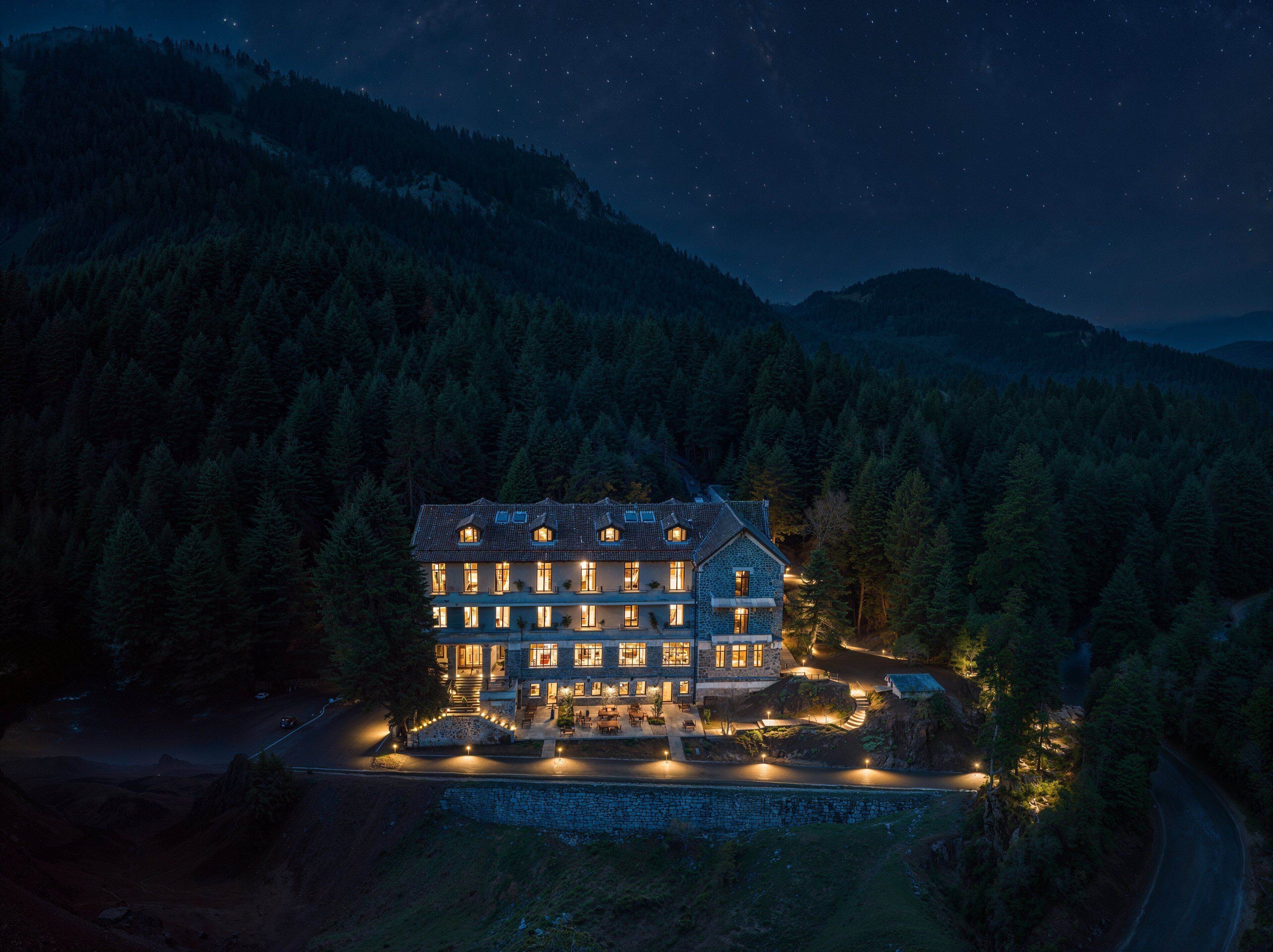 A large, multi-story building with a stone facade, surrounded by dense forest and mountains.
