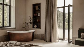 A modern bathroom with a freestanding bathtub, a bookshelf, and a view of a forest through large windows.