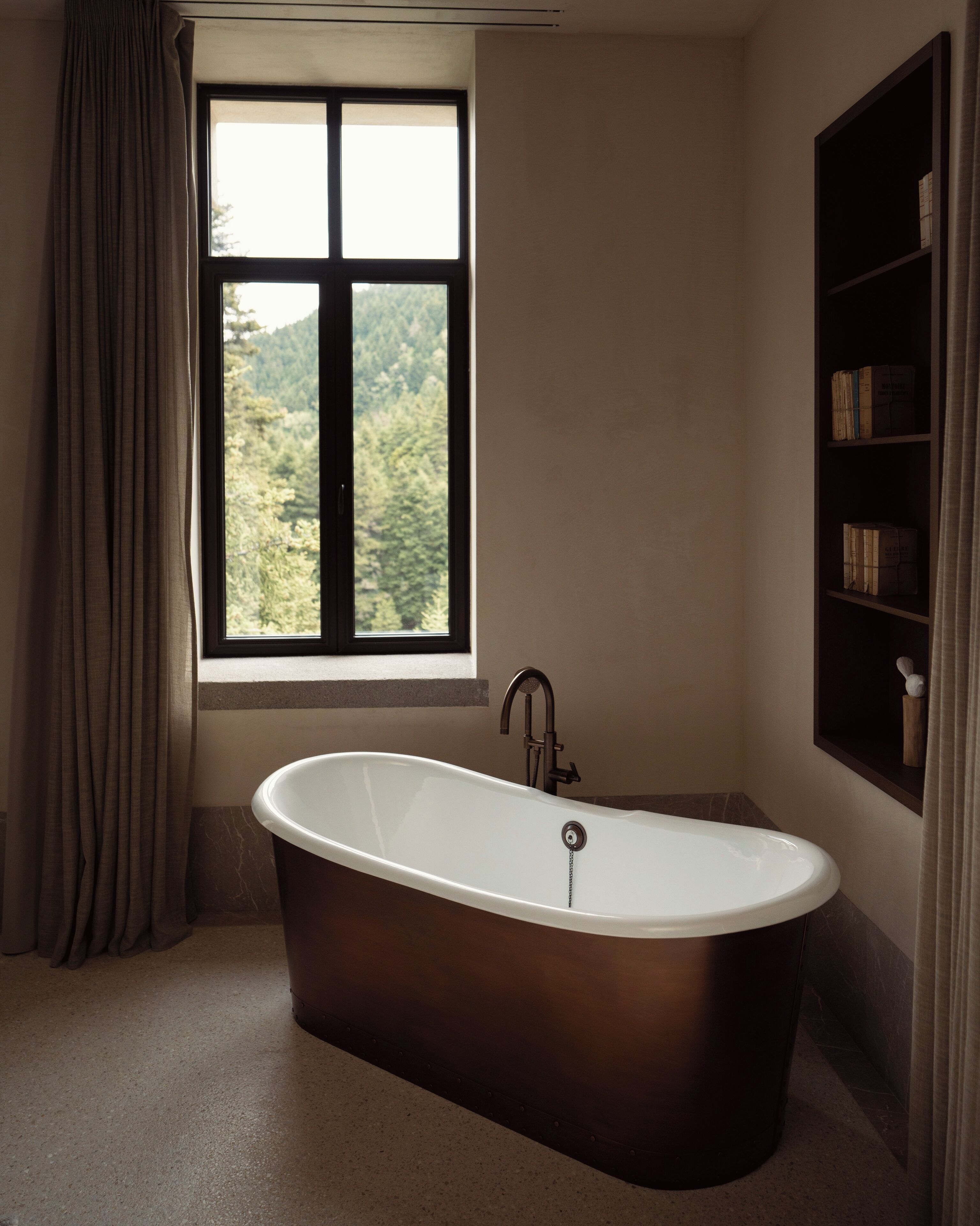 A bathroom with a freestanding bathtub, a window with a view of trees, and a shelf with books.