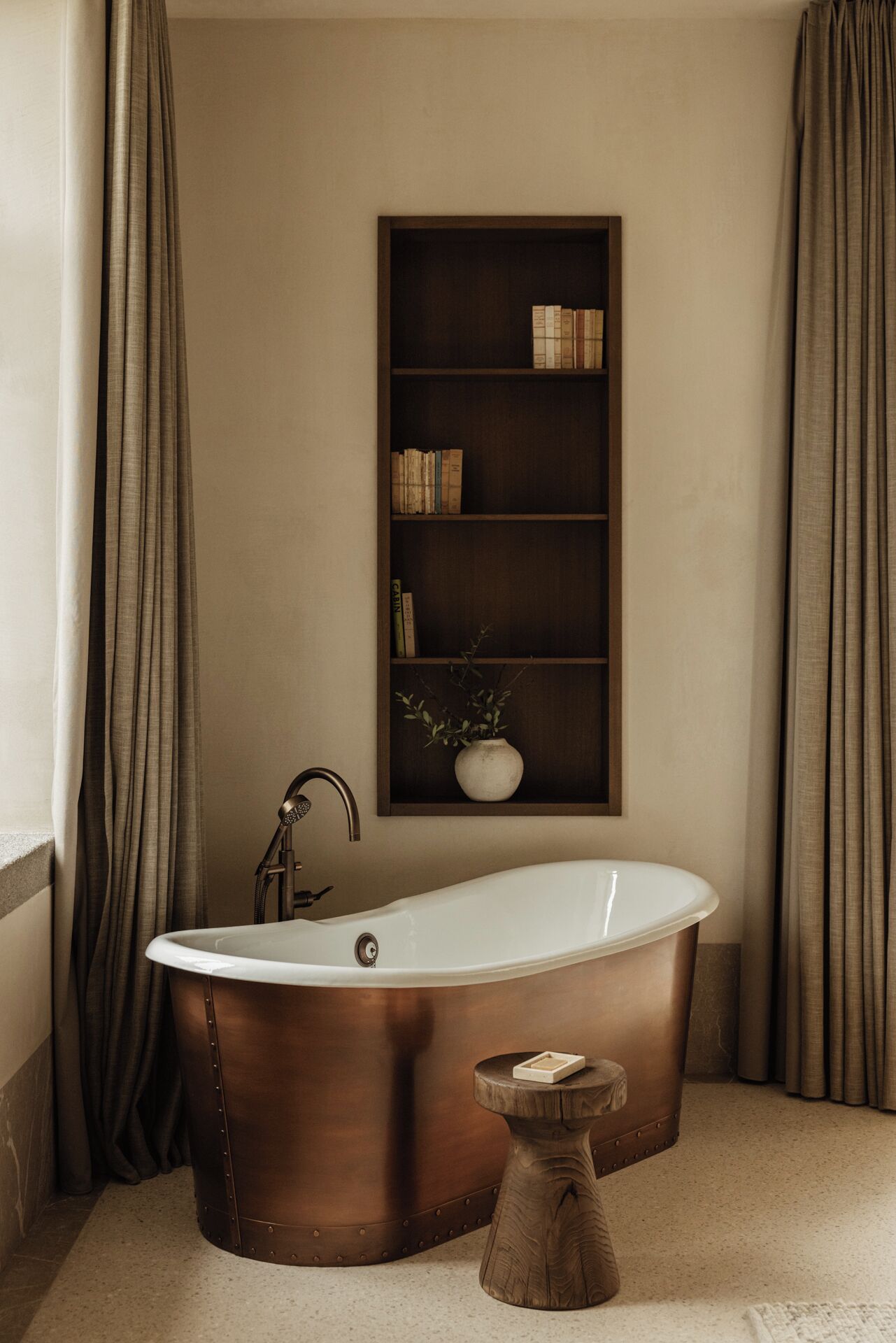 A bathroom with a freestanding bathtub, a wooden shelf with books and a vase, and a small wooden stool.