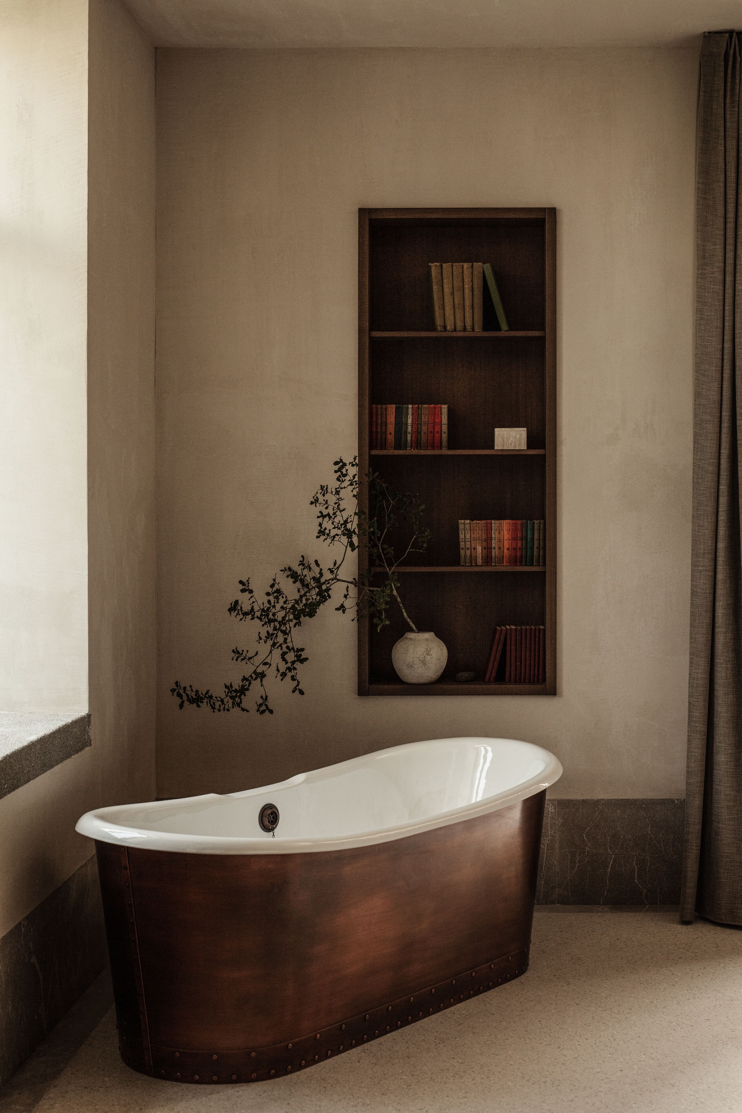 A bathroom with a freestanding bathtub, a bookshelf with books and decorative items, and a potted plant.
